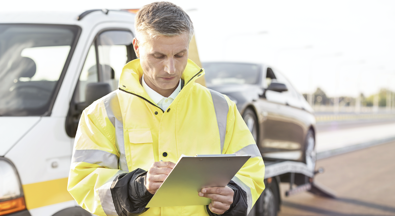 A man starts anding on the side of the road next to a tow truck taking notes.