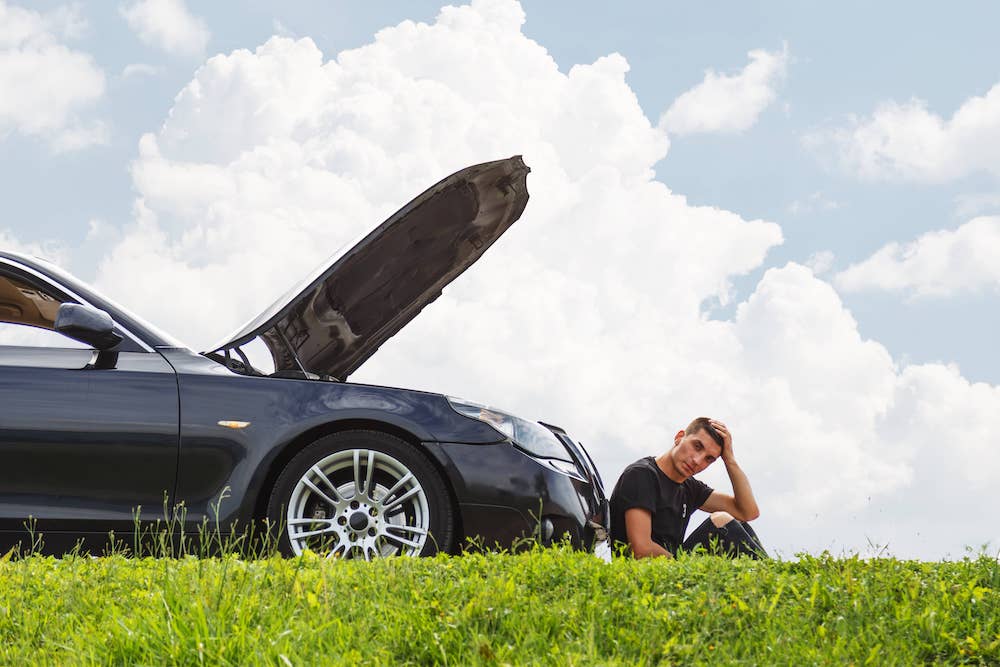 A young man leading up against his broken down car next to green grass while he figures out what to do.