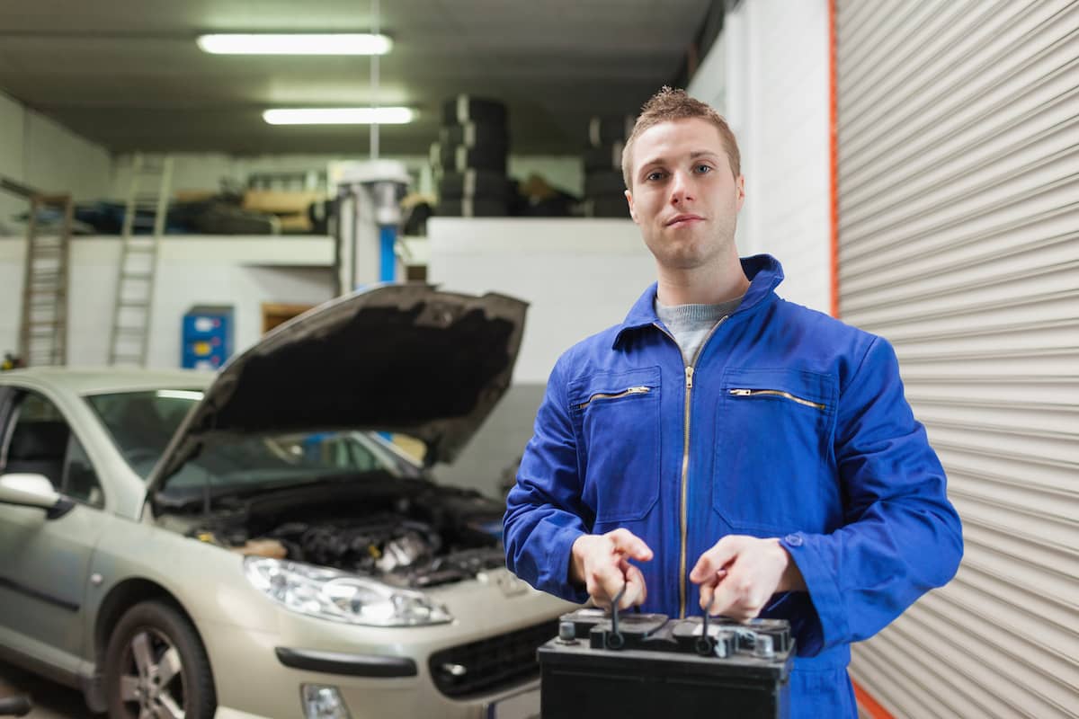 A mechanic standing in a garage holding a car battery.